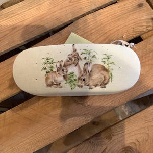 May include: An oval-shaped eyeglasses case with a beige linen texture. The case features a watercolor illustration of three brown rabbits with green foliage. The case has a white ribbon attached to the side.