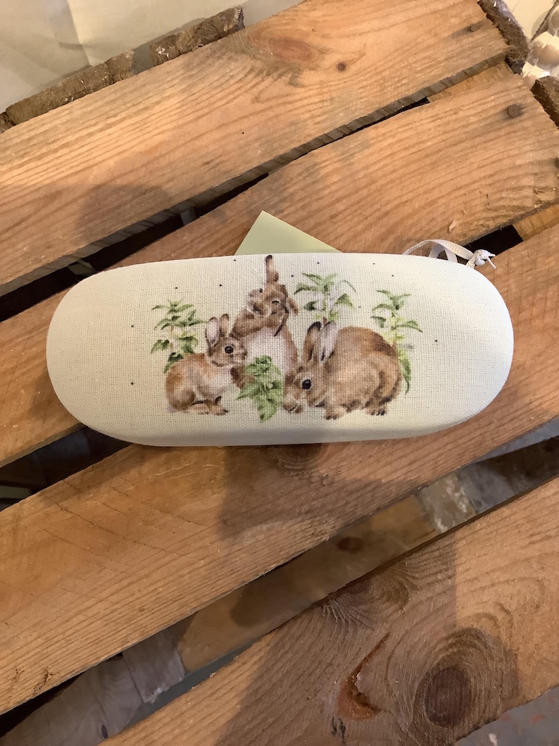 May include: An oval-shaped eyeglasses case with a beige linen texture. The case features a watercolor illustration of three brown rabbits with green foliage. The case has a white ribbon attached to the side.