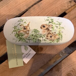 May include: An oval-shaped glasses case with a light green background. The case features a watercolor illustration of three brown bunnies amidst green foliage. A tag reads "100% Recycled Cotton."