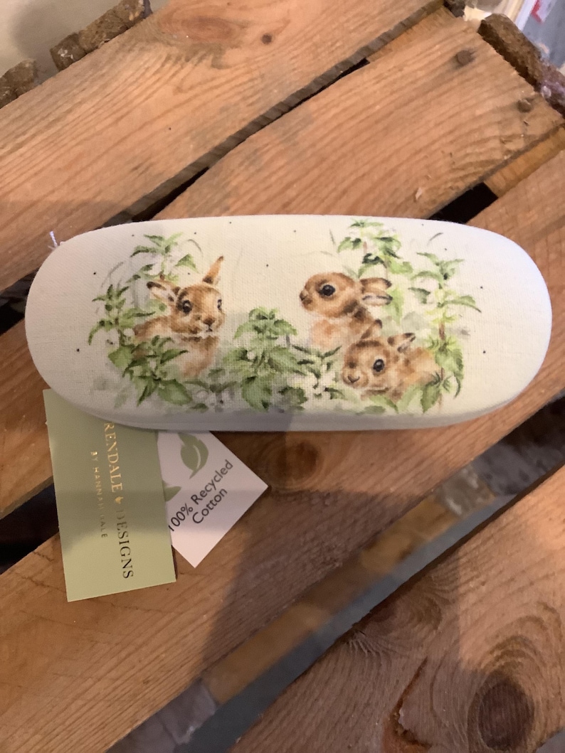 May include: An oval-shaped glasses case with a light green background. The case features a watercolor illustration of three brown bunnies amidst green foliage. A tag reads "100% Recycled Cotton."