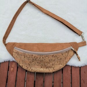 Può includere: Un marsupio a mezzaluna con una sezione superiore in finta pelle scamosciata color marrone chiaro e una sezione inferiore in sughero naturale con un motivo a macchie marrone scuro. La borsa presenta una chiusura a zip beige e una tracolla.