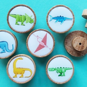 Puede incluir: Conjunto de siete pomos de madera con ilustraciones de dinosaurios coloridos. Los pomos son redondos y tienen un fondo blanco con una imagen de dinosaurio en cada uno. Los dinosaurios incluyen un triceratops verde, un estegosaurio azul, un pterodáctilo rosa, un braquiosaurio amarillo, un tiranosaurio rex verde y un plesiosaurio azul. Los pomos son perfectos para agregar un toque divertido y caprichoso a cualquier habitación.