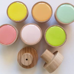 Könnte beinhalten: Eine Sammlung runder Holzknöpfe mit glatten, glänzenden Harzeinlagen in den Farben Gelb, Orange, Mintgrün, Koralle, Hellrosa und Limettengrün, wobei zwei Knöpfe von der Seite gezeigt werden, um die natürliche Holzmaserung zu enthüllen.