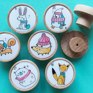 May include: A selection of wooden cabinet knobs with charming winter animal illustrations. Each knob showcases a different animal, including a rabbit, penguin, squirrel, hedgehog, bear, and fox, all adorned with winter attire. The knobs have a natural wood finish, set against a teal backdrop.