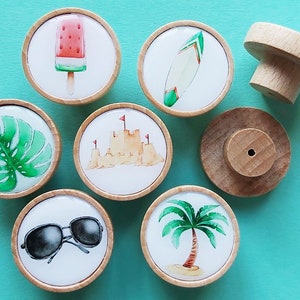 May include: Set of 7 wooden knobs with colourful summer-themed illustrations. The knobs feature images of a watermelon popsicle, a surfboard, a palm tree, sunglasses, a sandcastle, a monstera leaf, and a plain wooden knob.