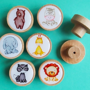 Könnte beinhalten: Eine Sammlung von Holzschrankknöpfen mit entzückenden Cartoon-Tieren: ein Bär, ein Zebra, ein Elefant, eine Giraffe, ein Waschbär und ein Löwe. Jeder Knopf hat eine runde, hellfarbene Holzbasis mit einer farbenfrohen Tierillustration.