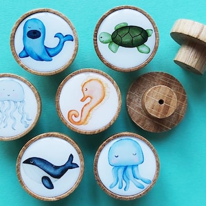 Op de afbeelding: Een verzameling houten kastknoppen met illustraties van zeedieren. Elke knop toont een ander zeedier, waaronder een zeehond, schildpad, kwal, zeepaardje, walvis en een blauwe kwal. De knoppen hebben een natuurlijke houten afwerking.
