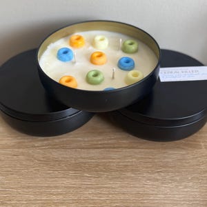 May include: A black candle tin with a cream-colored wax base, decorated with colorful cereal pieces. The candle has three wicks and the text "Cereal Killer" on the lid. The tin is sitting on a wooden surface.