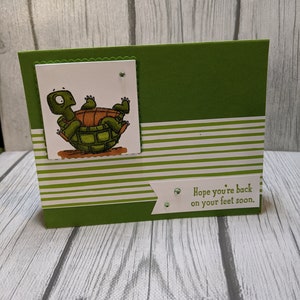 May include: A handmade get-well card with a green and white striped design. The card features a cartoon turtle illustration and the text "Hope you're back on your feet soon."
