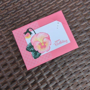 May include: A pink birthday card with a floral design. The card features a large, detailed flower with pink and yellow petals, green leaves, and the words "Happy Birthday." The card is inside a pink envelope.