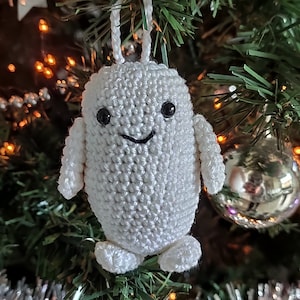 Op de afbeelding: Witte gehaakte sneeuwman-versiering met zwarte ogen en een glimlachende mond die aan een kerstboom hangt.