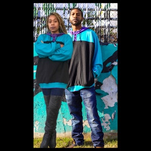 Op de afbeelding: Twee mensen dragen blauwe en zwarte sweatshirts met een turquoise rand. De vrouw draagt ​​zwarte leggings en zwarte laarzen. De man draagt ​​een blauwe jeans. Ze staan ​​voor een blauwe en groene muur met een gaashek erachter.
