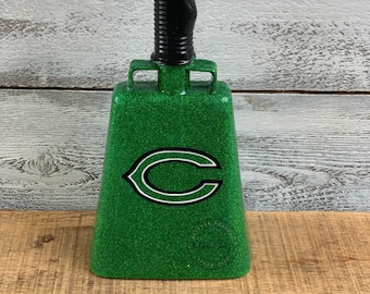 Op de afbeelding: Groene glitter-koeienbel met een zwarte handgreep en een wit "C" logo. De koeienbel heeft een kleine sticker op de bodem.
