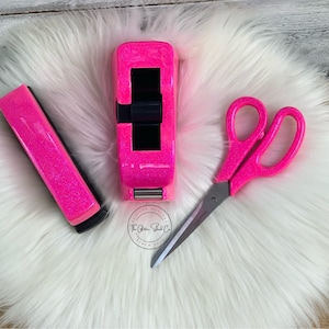May include: Pink glitter tape dispenser, pink glitter stapler, and pink glitter scissors on a white fluffy surface. The Glitter Shack Co. is printed on the surface.