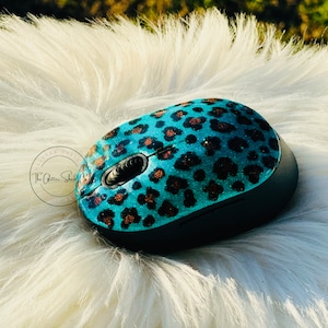 May include: A wireless computer mouse with a teal leopard print design. The mouse has a black base and features a glittery finish. The text "The Glitter Shed Co" is visible on the left side of the mouse.