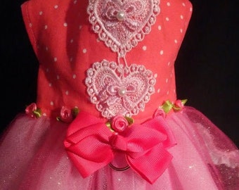 May include: A pink and red dog dress with a tulle skirt and lace trim. The dress has two white lace hearts and a pink bow.