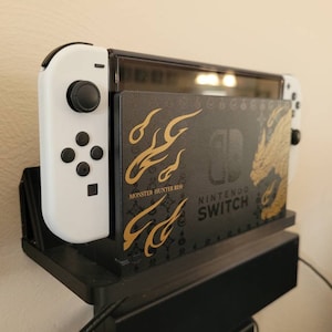 May include: A white Nintendo Switch console with a black and gold Monster Hunter Rise game case. The console is in a black stand with a black and gold design.