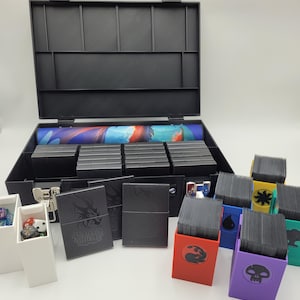 May include: A black plastic carrying case with an open lid, containing card storage boxes, a rolled-up play mat, and various card holders in different colors. Several card holders are visible, each with a different symbol.