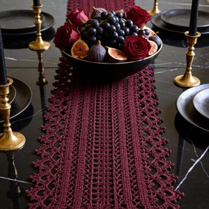 Pode incluir: Um caminho de mesa de crochê bordô com uma borda decorativa, exibido em uma mesa de mármore preto. A mesa está posta com pratos pretos, castiçais dourados e uma tigela de uvas, figos e rosas. Há velas pretas nos castiçais.