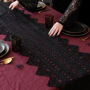 May include: A burgundy tablecloth is set with black plates, gold cutlery, and a black lace runner. Black candles and a decorative raven are on the mantelpiece. The table is set for a gothic-themed dinner party.