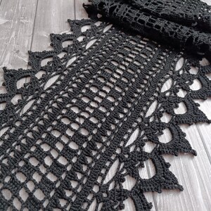 Peut inclure: Un chemin de table en crochet noir présentant un motif central en grille composé de petites ouvertures carrées. Les bords du chemin de table sont finis avec une bordure décorative en forme de pointes et de découpes circulaires texturées.