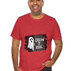 Peut inclure: T-shirt rouge avec un motif de fantôme de dessin animé regardant derrière un mur de briques. Le texte "CREEPIN' IT REAL" est affiché sur le mur. Le t-shirt est de couleur rouge uni.