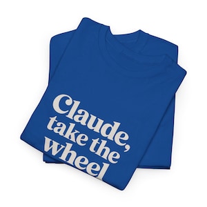 Op de afbeelding: Twee gevouwen koningsblauwe t-shirts. Het bovenste shirt heeft de zin "Claude, take the wheel" in witte, vette, schreefloze lettertype. De shirts zijn gemaakt van een zacht materiaal en zijn netjes opgevouwen.