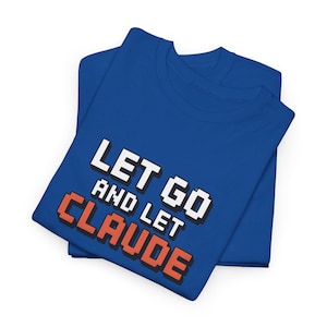 Puede incluir: Dos camisetas azules dobladas y apiladas. La camiseta superior presenta el texto "LET GO AND LET CLAUDE" en fuente pixelada blanca y naranja. Las camisetas están hechas de un material suave.
