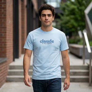 Puede incluir: Una camiseta de manga corta y cuello redondo de color azul claro con el texto azul oscuro "claude is my copilot" impreso en una fuente redondeada y negrita en el centro del pecho. La prenda está hecha de tela de color sólido.