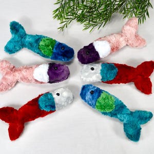 May include: Six plush fish toys in various colors, including teal, pink, red, and white. Each fish has a different color combination, with a fluffy texture and a small black eye. The toys are arranged on a white surface.
