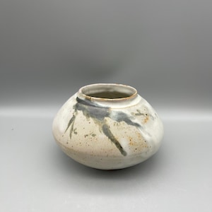 以下が含まれることがあります： 丸みを帯びたボディと広い開口部を持つ、手作りのセラミック花瓶。 花瓶は主に白で、斑点模様と濃いグレーとグリーンの抽象的な筆致が特徴です。 内側は暗い色合いで、素朴で有機的な美しさを持っています。