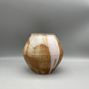 Może przedstawiać: Ręcznie wykonany ceramiczny wazon o zaokrąglonym kształcie. Wazon ma nakrapianą brązową podstawę z białą i różową glazurą, która spływa po boku. Obręcz jest w jasnobeżowym kolorze.