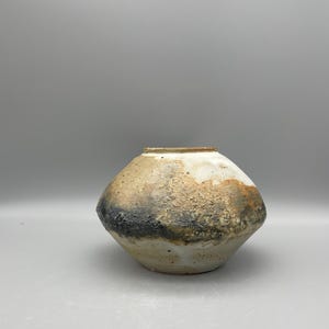 Może przedstawiać: Ręcznie wykonany ceramiczny wazon o unikalnej, teksturowanej powierzchni. Wazon ma zaokrąglony, asymetryczny kształt z szerokim otworem. Paleta kolorów obejmuje odcienie bieli, beżu, czerni i brązu, tworząc rustykalną, ziemistą estetykę. Wazon nadaje się do dekoracji wnętrz.