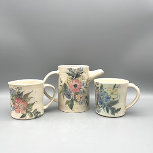May include: A ceramic tea set featuring a teapot and two mugs. The set is off-white with hand-painted floral designs in pink, blue, and yellow. The teapot has a spout and handle, while the mugs have curved handles.