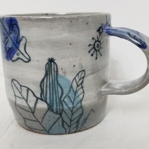 May include: A white ceramic mug with a blue and grey hand-painted design of leaves and a cactus. The mug has a rounded shape and a handle.