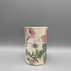 May include: A white ceramic vase with a floral pattern. The vase is decorated with pink and white flowers and green leaves.
