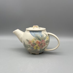 Könnte beinhalten: Eine kleine weiße Teekanne mit Blumenmuster. Die Teekanne hat einen runden Korpus, einen Ausguss, einen Henkel und einen Deckel. Die Blumen sind in Rosa-, Gelb- und Blautönen bemalt.