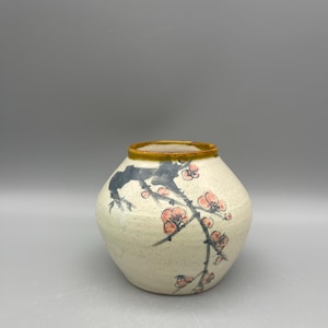 May include: A small, round, white ceramic vase with a brown rim and a painted design of a branch with pink flowers.