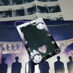 May include: A rectangular ID holder with a black interior and a white border featuring a cartoon bunny face with a pink bow. The holder has a heart-shaped cutout and a purple beaded chain.