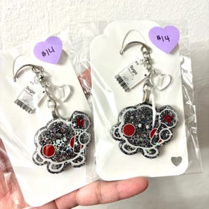 May include: Two identical keychains featuring a black, glittery, cloud-shaped character with red circular cheeks and a small red symbol. Each keychain includes a silver metal ring, a small clear heart charm, and a rectangular tag labeled BTS Suga.
