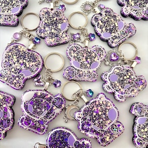 May include: A collection of purple and black glitter resin keychains featuring a cartoon koala character holding a microphone. Each keychain includes a silver metal ring and various small charms like stars, hearts, and moons attached to the top.