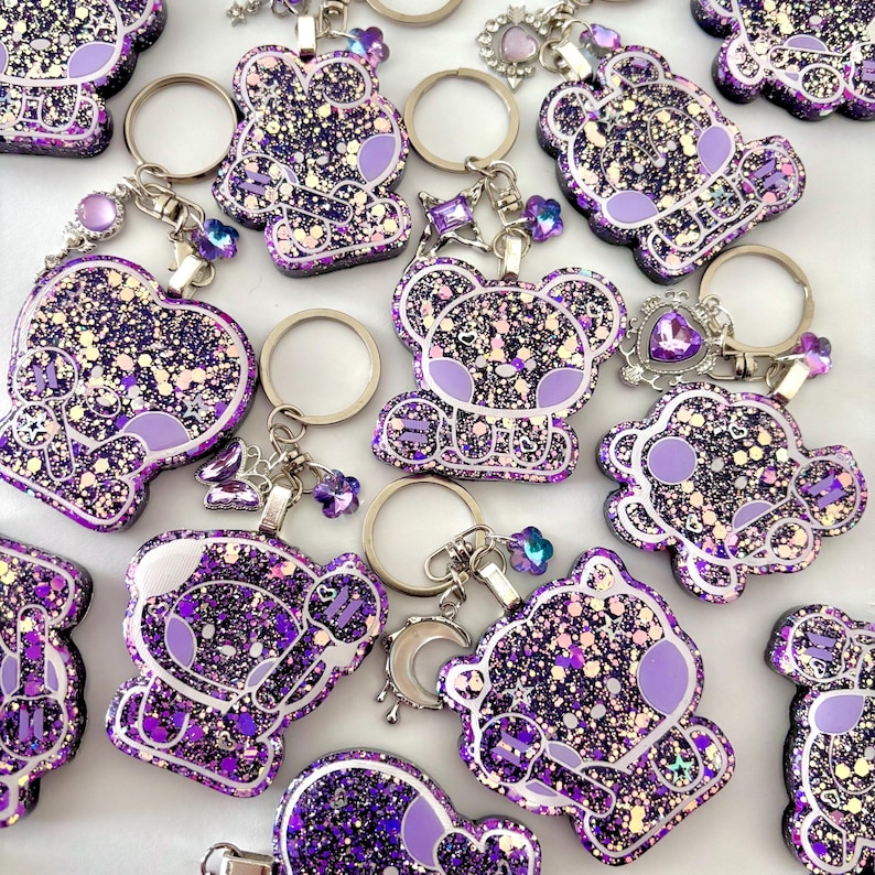 May include: A collection of purple and black glitter resin keychains featuring a cartoon koala character holding a microphone. Each keychain includes a silver metal ring and various small charms like stars, hearts, and moons attached to the top.