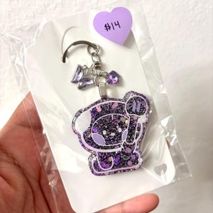 May include: A purple and black glittery keychain featuring a cartoon character holding a light stick. The keychain includes a silver metal ring, a small purple butterfly charm, and a small purple flower charm, all packaged in a clear plastic bag.