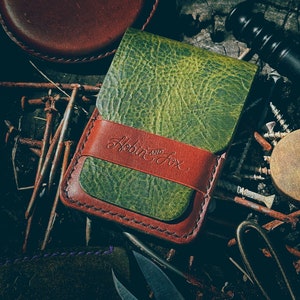 May include: A green leather wallet with a red leather flap and the text "Hobbit and Fox" embossed on the flap. The wallet is lying on a surface with rusty nails and other metal objects.