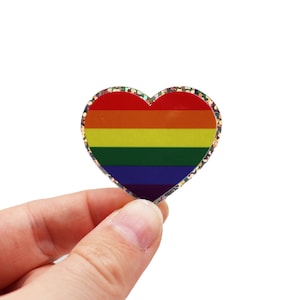 Könnte beinhalten: Ein herzförmiger Anstecker mit Regenbogenflaggen-Design. Der Anstecker hat horizontale Streifen in Rot, Orange, Gelb, Grün, Blau und Lila. Die Ränder sind mit Glitzer verziert. Der Anstecker wird von einer Hand vor weißem Hintergrund gehalten.