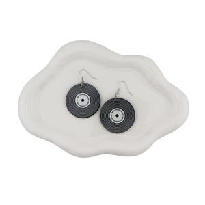 May include: A pair of black and white vinyl record earrings with silver hooks. The earrings are circular with concentric lines and a white center. The text "HOPE LOVE RECORDS" is printed on the white center. The earrings are displayed on a white, cloud-shaped dish.
