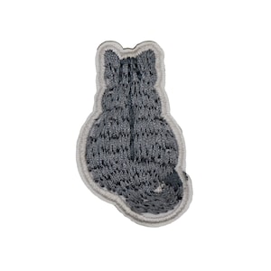 Peut inclure: Un écusson brodé en forme de chat gris, vu de dos, avec une texture duveteuse. L'écusson a une bordure gris clair et est conçu pour être cousu sur du tissu. La queue du chat est légèrement recourbée vers la droite.