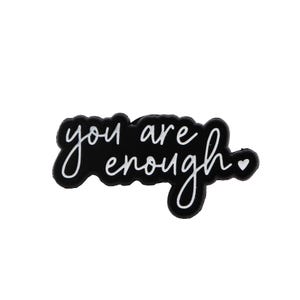 Könnte beinhalten: Eine schwarze Anstecknadel mit den Worten "you are enough" in weißer Schreibschrift. Die Anstecknadel hat eine abgerundete rechteckige Form mit einem kleinen Herz am Ende des Wortes "enough".