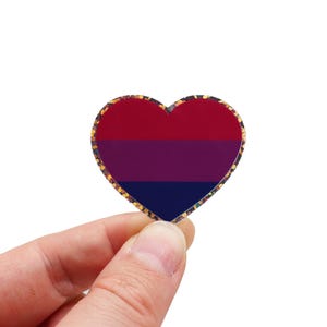 Könnte beinhalten: Ein herzförmiger Anstecker mit einem goldglitzernden Rand. Der Anstecker zeigt die bisexuelle Pride-Flagge: drei horizontale Streifen in Rot, Lila und Marineblau. Der Anstecker wird in einer Hand vor weißem Hintergrund gehalten.