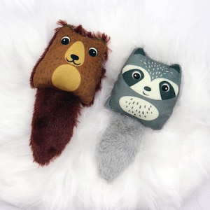Può includere: Due peluche di animali: un orso marrone e un procione grigio. L'orso ha un corpo marrone, una faccia beige e una coda rosso scuro. Il procione ha un corpo grigio, una faccia bianca e una coda grigia. Entrambi i giocattoli hanno dettagli ricamati.